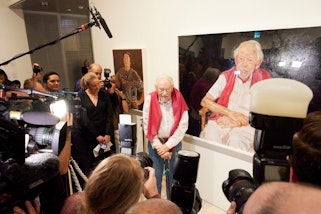 In a gallery space surrounded by media with cameras, an elderly man wearing a white shirt with a pink jumper over his shoulders stands in front of a painting of him seated in similar clothes. 