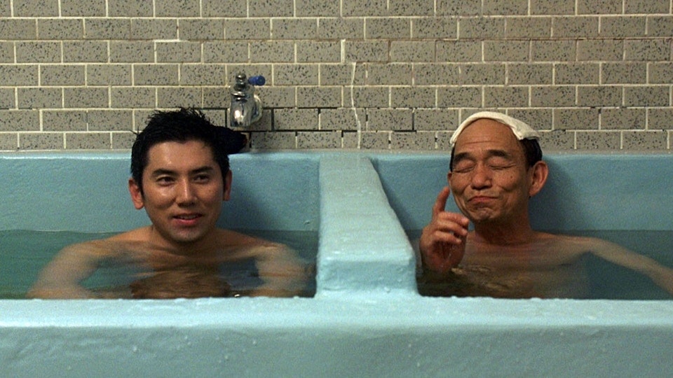 Two people with light brown skin sitting in a partitioned bath. One of them has a towel over their head.