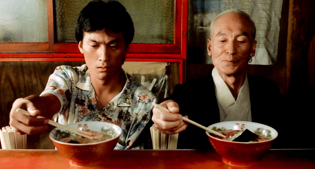 Two people with light brown skin placing chopsticks into bowls of ramen. The person on the left is frowning.