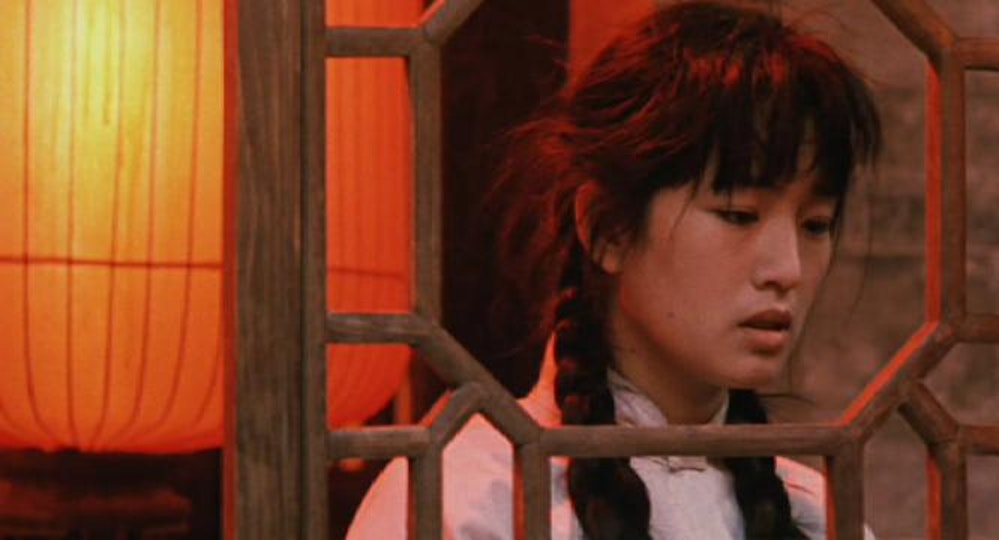 A young person with black hair in plaits looks through an ornate wooden frame. Behind the person is a red lantern.