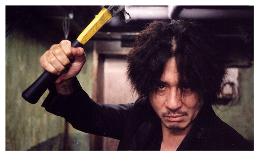 A person with shaggy black hair and a oustache raises a hammer.