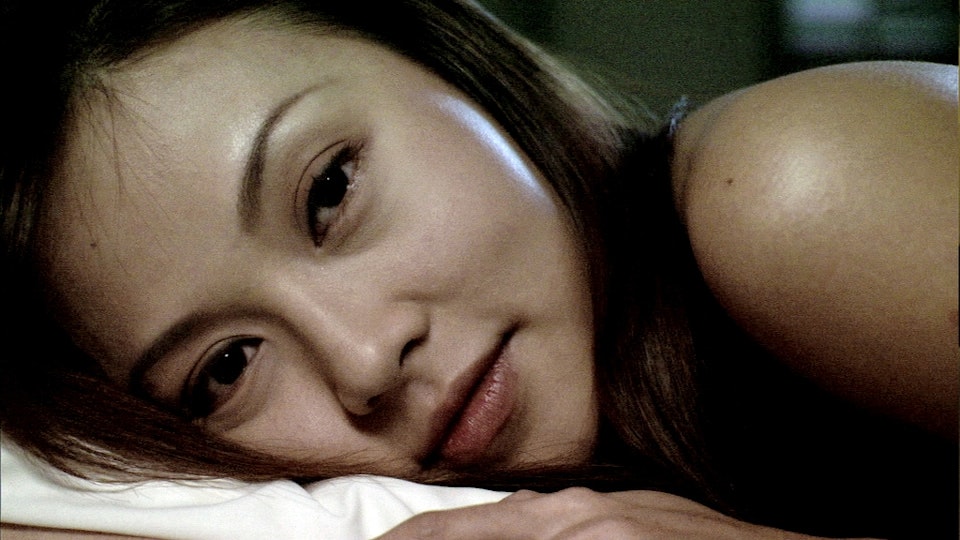 A person with light skin and long black hair is lying on a pillow, looking into the camera.