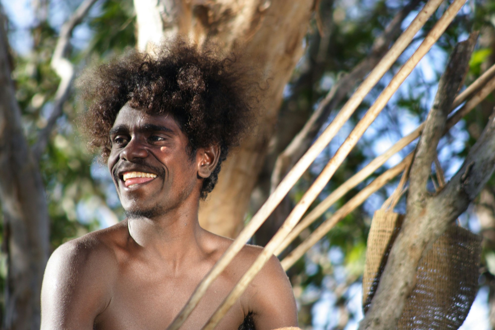 A person with dark brown skin smiles in front of a tree.
