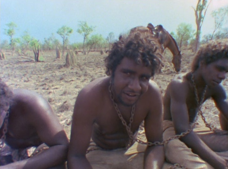 Three shirtless people with dark brown skin sit on the ground with chains around their necks.