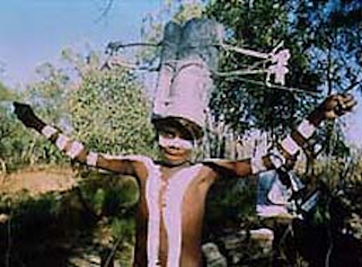A child with dark brown skin and ceremonial paint markings on their body smiles and stretches out their arms.