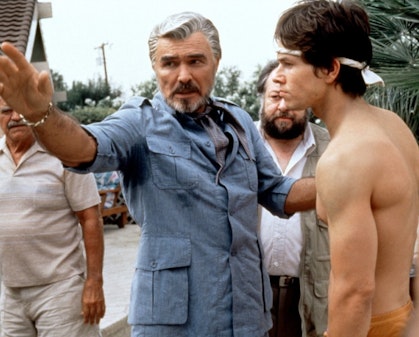A person with white hair and a blue suit has an arm on a shirtless person.