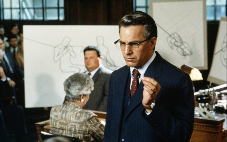 A person with light skin wearing glasses holds up their left hand. Behind them are large drawings.