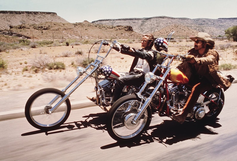 Two people driving motorcycles that are on a road next to plateaus.