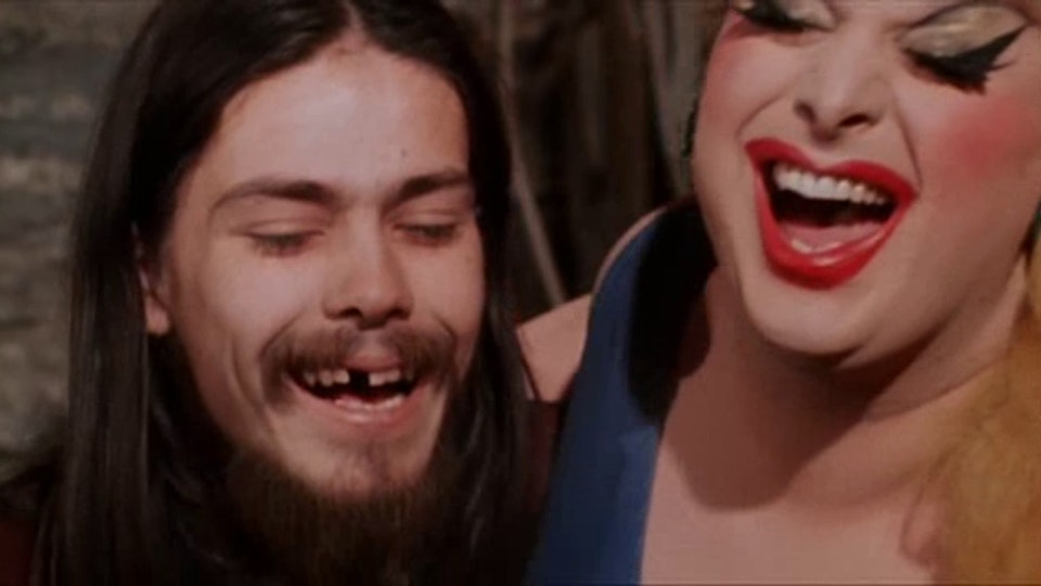 Two people with light skin laughing with their eyes closed. The person on the right has long fake eyelashes and red lipstick. The person on the left has a moustache and is missing one front tooth.