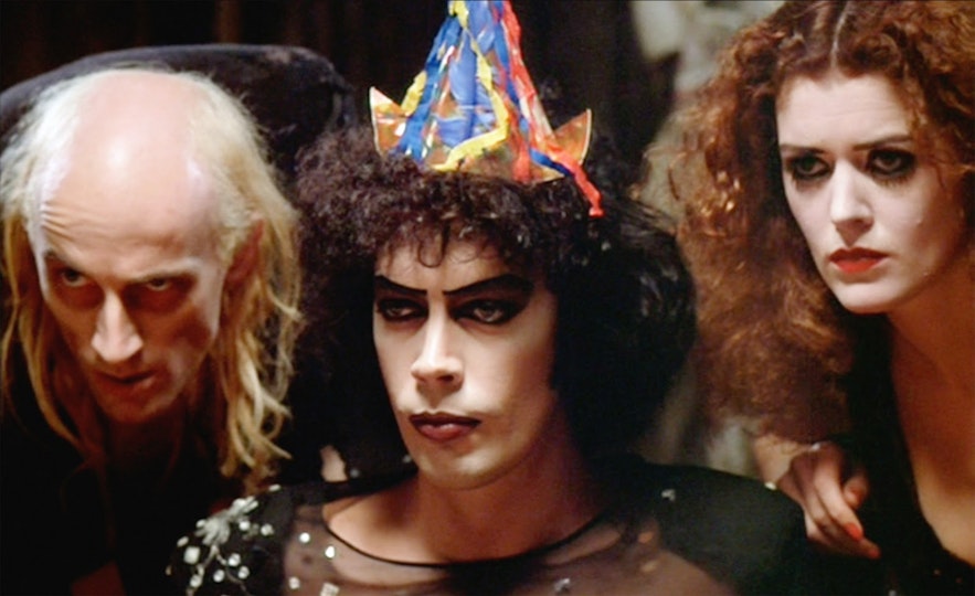Three people with pale faces look ahead of them. The person in the middle has dramatic drawn-on eyebrows and a party hat.