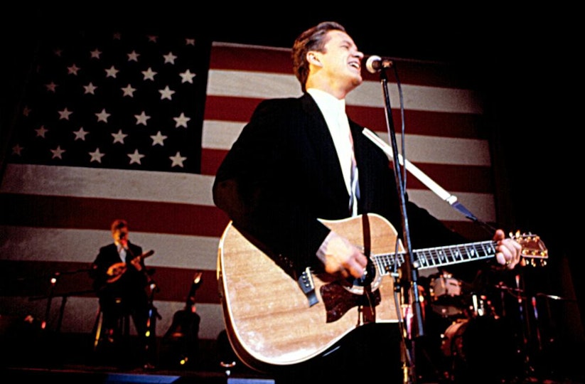 A person with light skin plays a guitar and sings into a microphone. Behind them is an enormous flag of the USA.