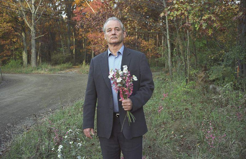 A man with light skin in a suit stands by the side of a forest road. He has a bunch of flowers in his hand.