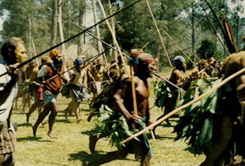 A group of people carrying wooden spears run across a grassy area.