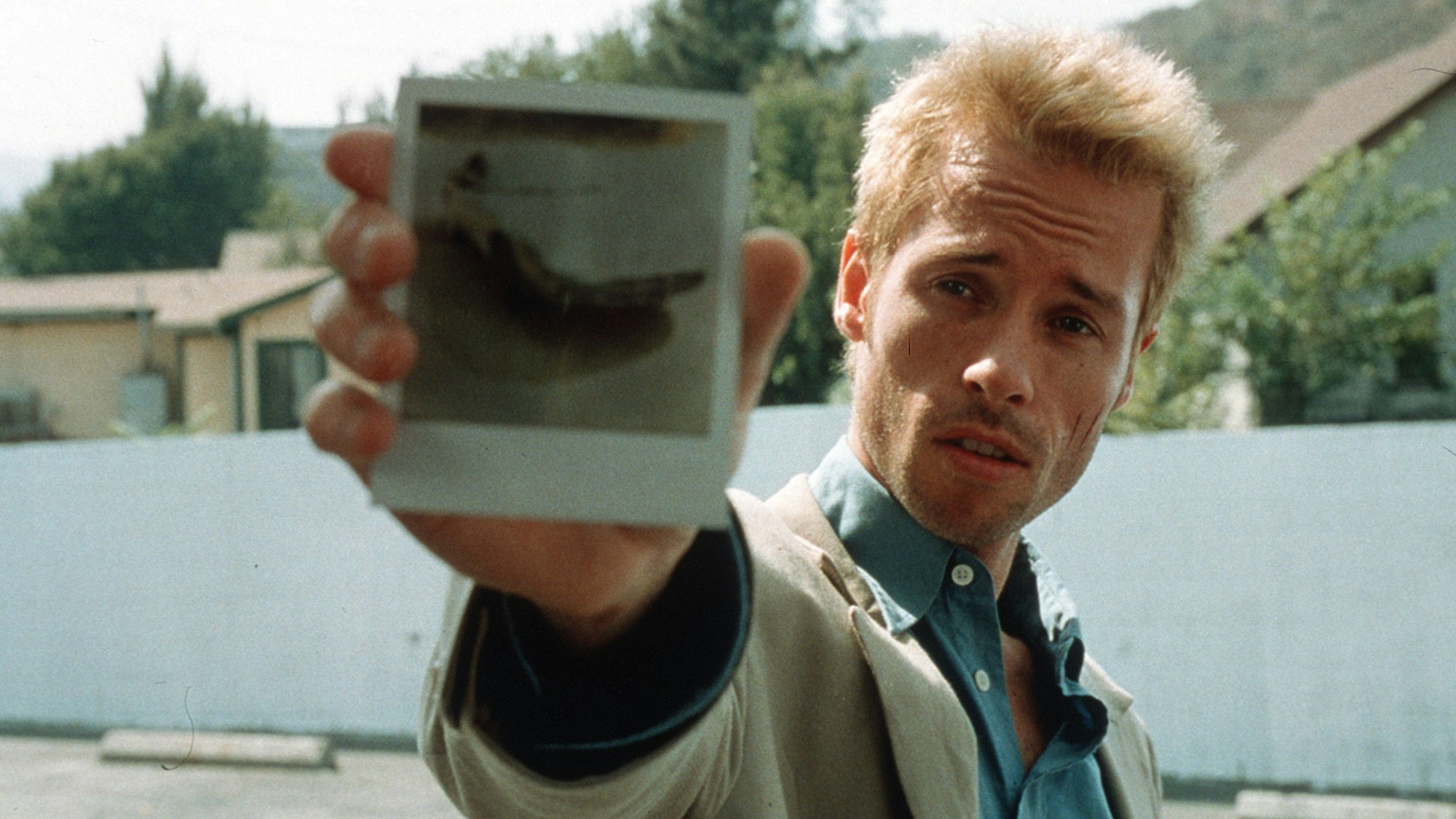 A man with bleached spiky hair and a gaunt face holds up a polaroid photo to the camera.