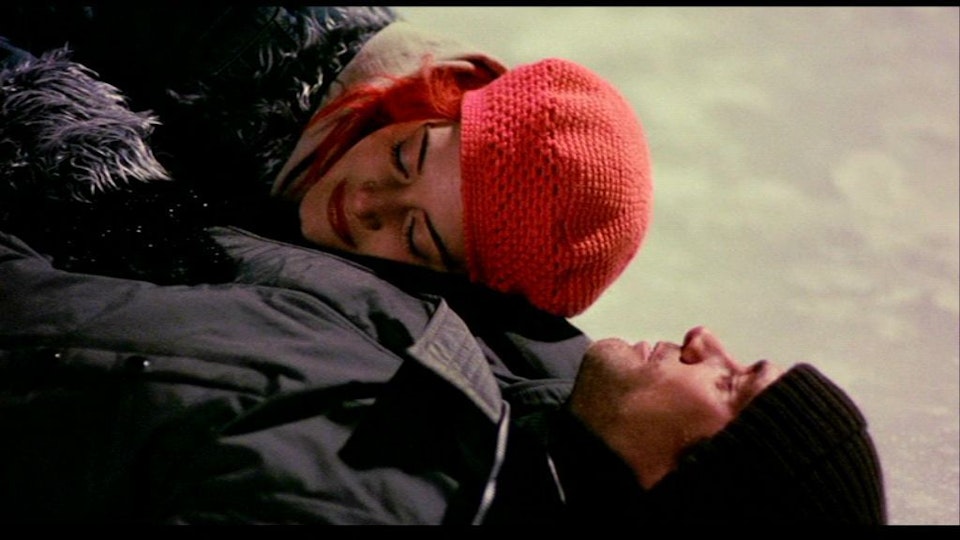 A woman with bright red hair and a red beanie lies on the shoulder of a man, who is lying on a frozen lake.