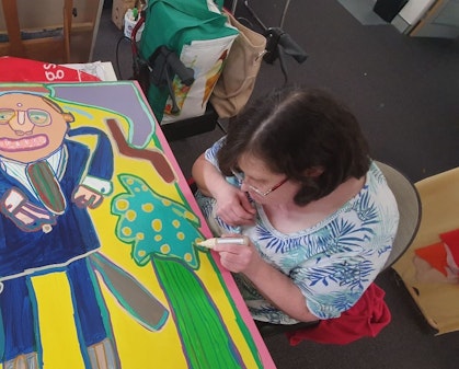 A person sitting in a chair works on a portrait painting, lying in front of them, of a person in a blue suit