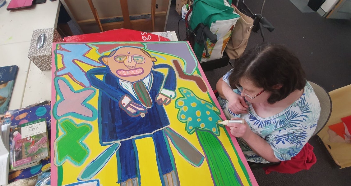 A person sitting in a chair works on a portrait painting, lying in front of them, of a person in a blue suit