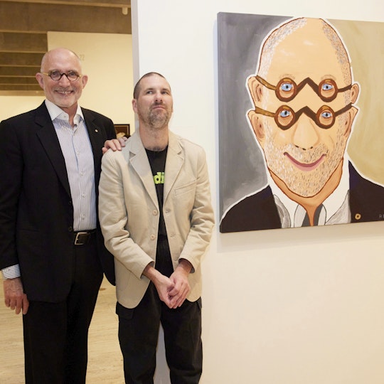 Two people, one with round glasses, stand in a gallery space next to a painting of the person with glasses. In the portrait, they are depicted with two pairs of glasses and two sets of ears.