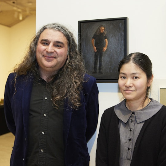 Two people stand in front of a small portrait painting. One of them is depicted in the portrait.