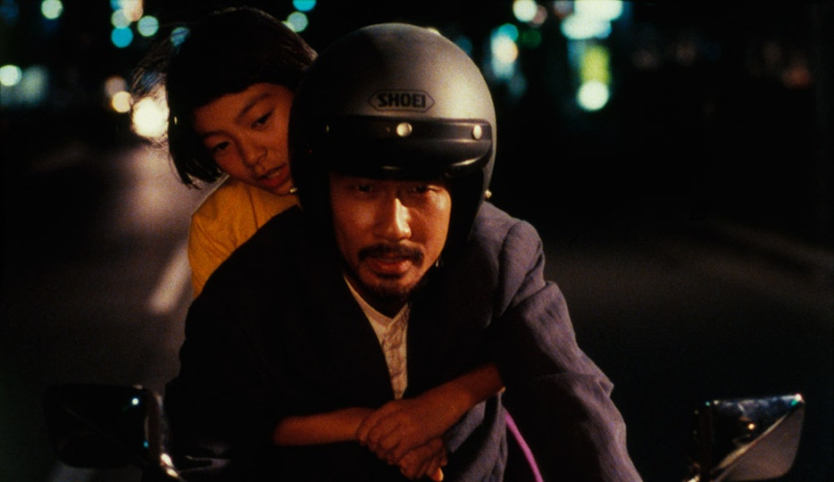 Two people riding on a motorbike at night. The person at the front has a moustache and a helmet. The person behind has short black hair.