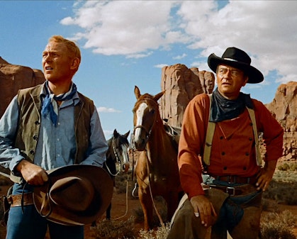 Two cowboys look in the distance. Behind them are two horses and a butte.