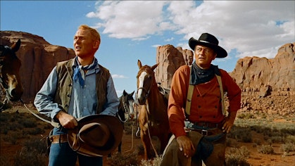 Two cowboys look in the distance. Behind them are two horses and a butte.