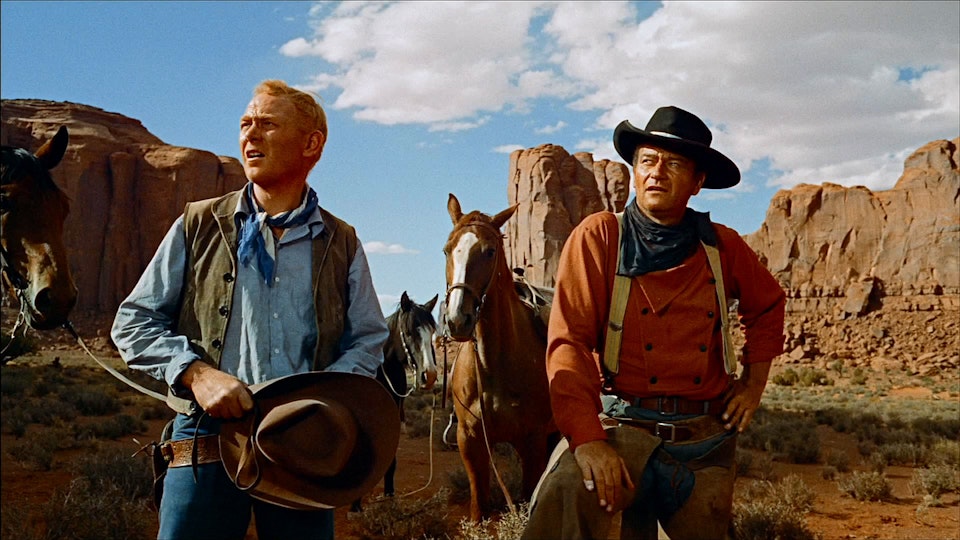 Two cowboys look in the distance. Behind them are two horses and a butte.