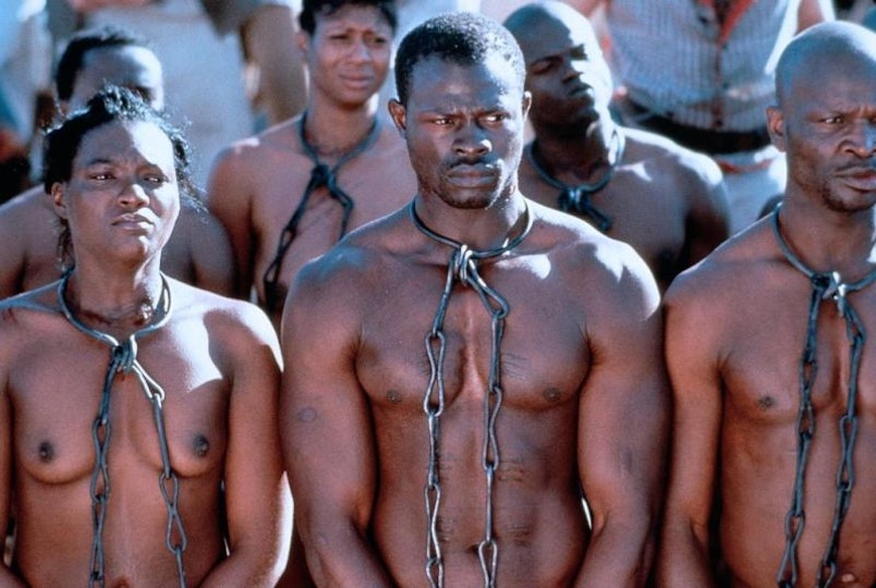 Six people with dark brown skin with chains around their necks. The sun is beating down on them.