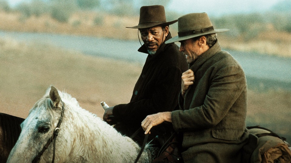 Two people wearing thick coats and fedoras are riding horses next to a road.