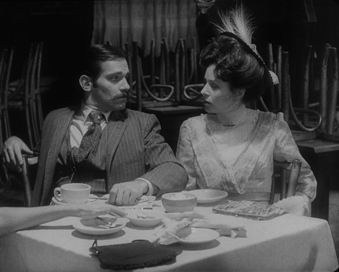 Two people seated at a table are facing each other. Behind them are stacks of upturned chairs.