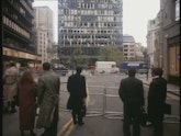 A group of people viewed from behind are standing in the middle of a barricaded road. In the ditance are tall buildings.