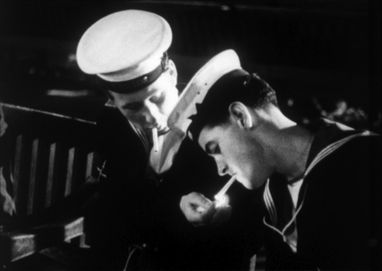 Two sailors bending their heads down to light cigarettes.