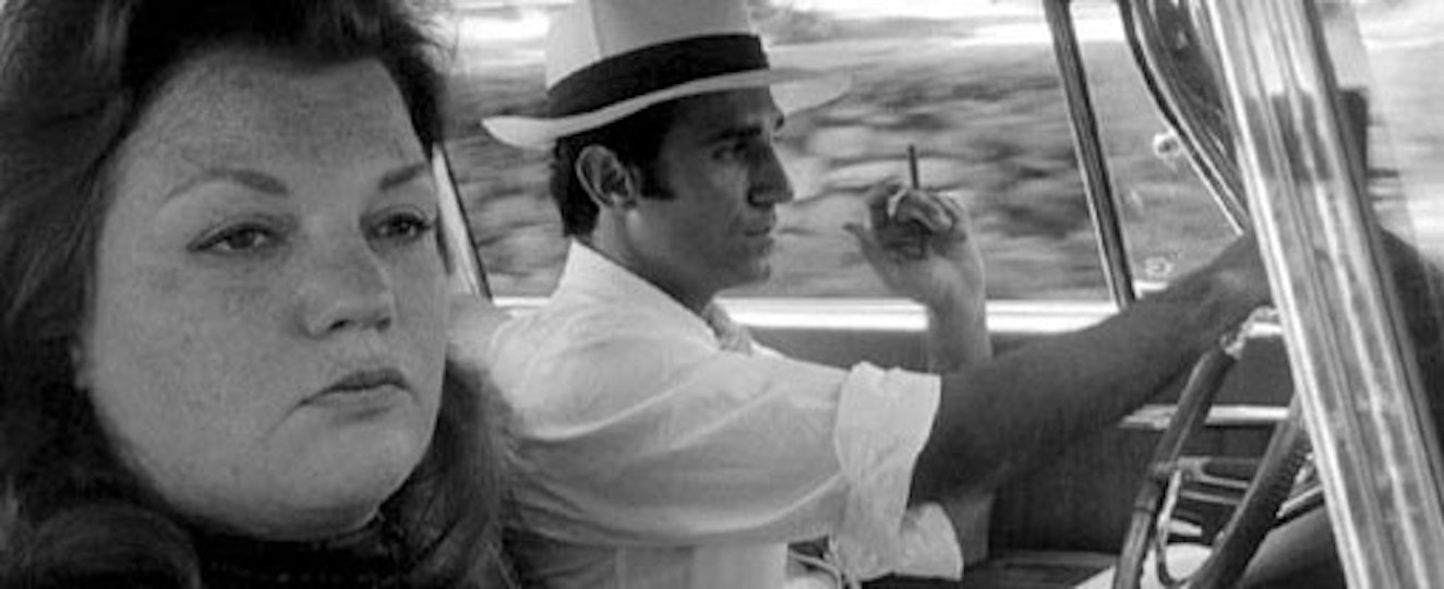 Two people seated in the front of a car. The driver wears a hat and holds a cigarette.
