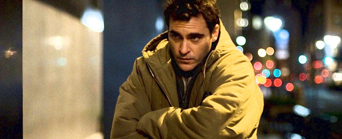 A person with short brown hair and light skin holds themselves in their thick coat as they walk down a street at night.