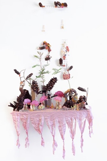 A hanging artwork consisting of objects such as plants and stones on a shelf covered with pink tasselled fabric.