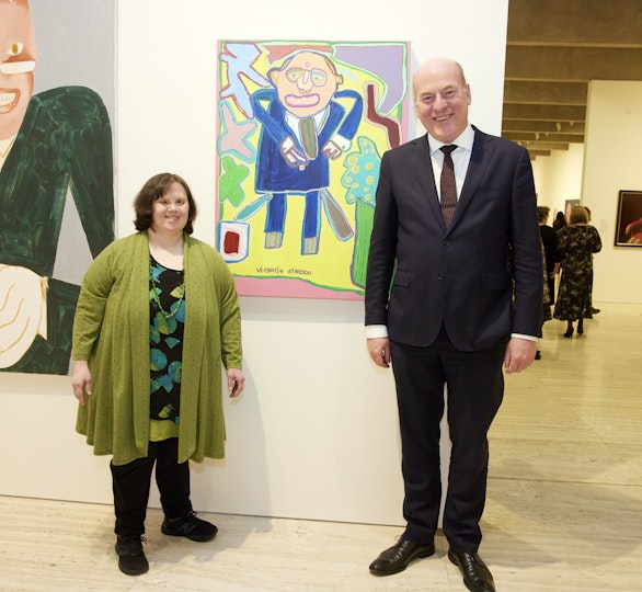 Two people stand either side of a portrait. Like in the painting, the person to its left is wearing a suit and tie and has a balding head and broad smile.