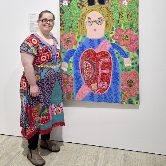 A person wearing glasses and a colourful dress stands next to a portrait of a bespectacled person with a background of flowers.