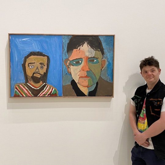 A person with short hair, wearing a t-shirt and vest, stands next to a painted portrait showing the heads and shoulders of two people.