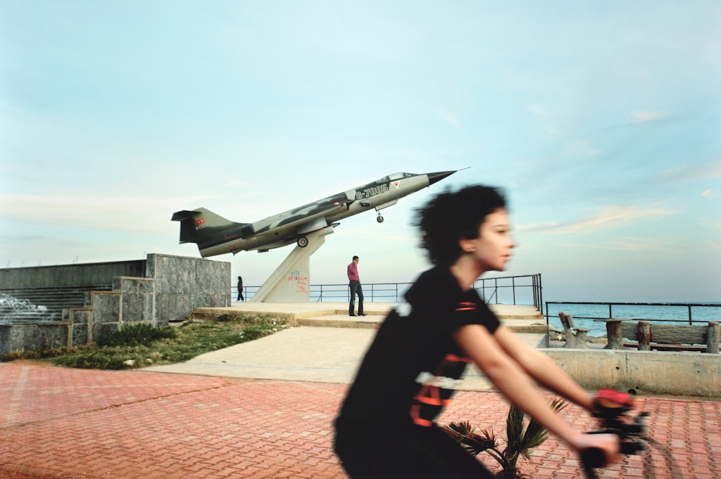 A person on a bike rides past a mounted fighter jet next to a fence, picnic table and ampitheatre on a hilltop. Two people walk near the plane.