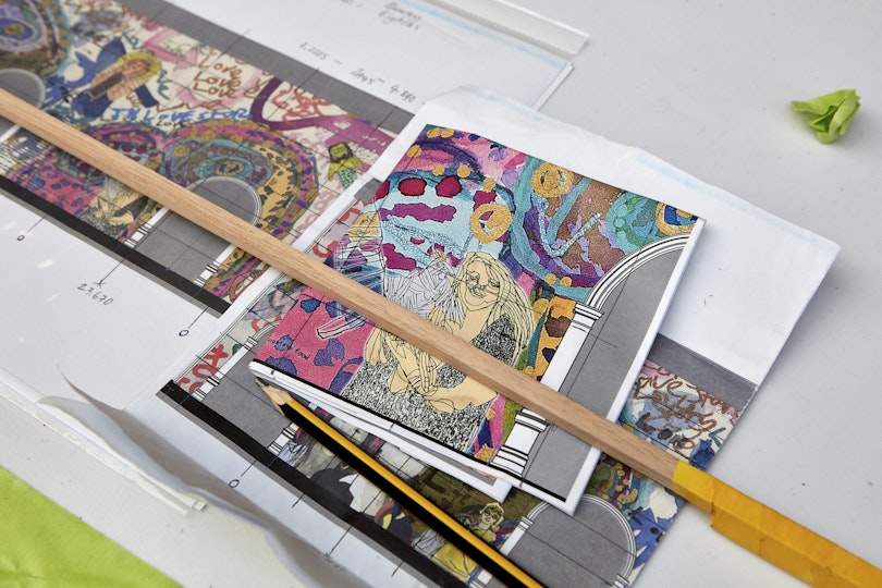 Pieces of paper with designs for a mural lie on a table with a pencil, wooden stick and plastic clip.