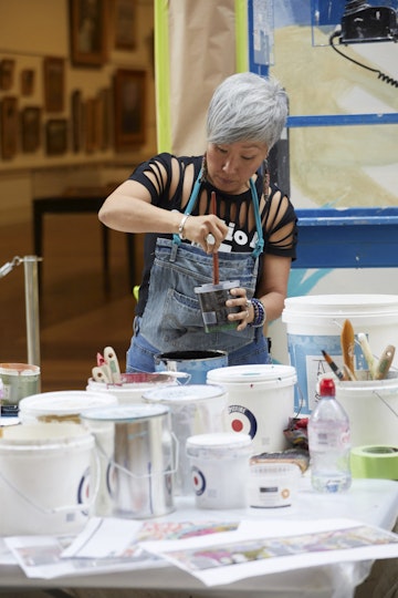A person mixes paint in a plastic tub while standing at a table full of paint buckets and other materials.