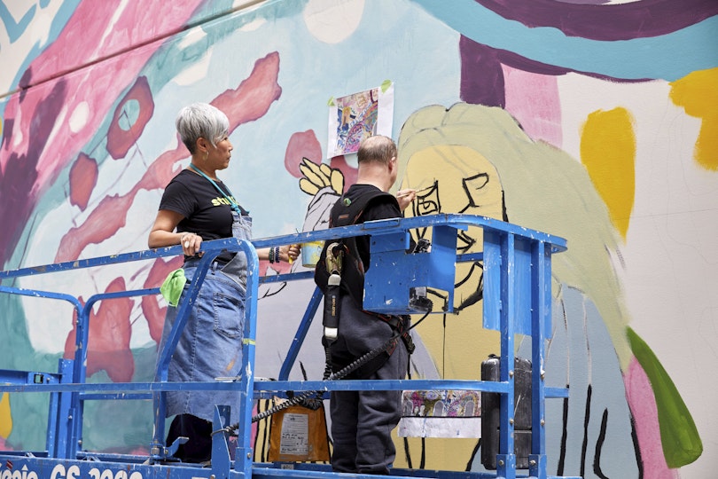 Two people stand on a platform in front of a mural. One is painting the wall while the other holds the paint container.