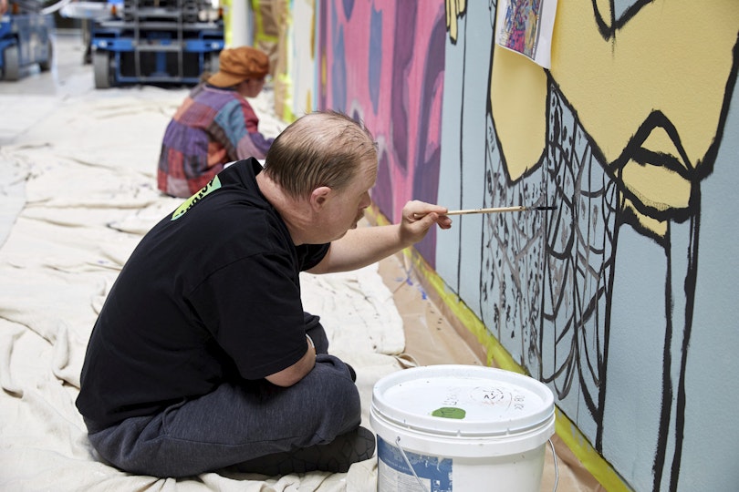 Two people sit cross-legged on a dropsheet on the floor while painting a mural.