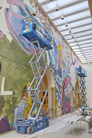 A person on a scissor lift paints the upper part of a wall, just under a high ceiling. Below them are work benches and a dropsheet.