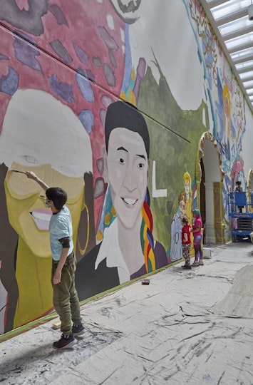 A person completes a portrait of a large head as part of a mural. Other artists work on different sections of the artwork.