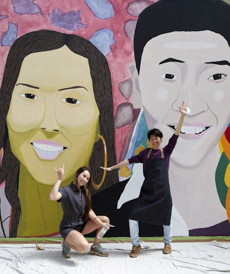 Two people pose in front of two large head-and-shoulder portraits on a wall.