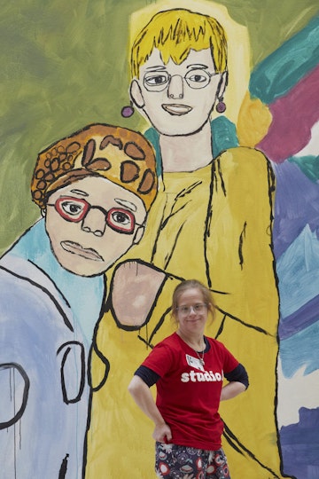 A person poses in front of a painting of two people on a wall.