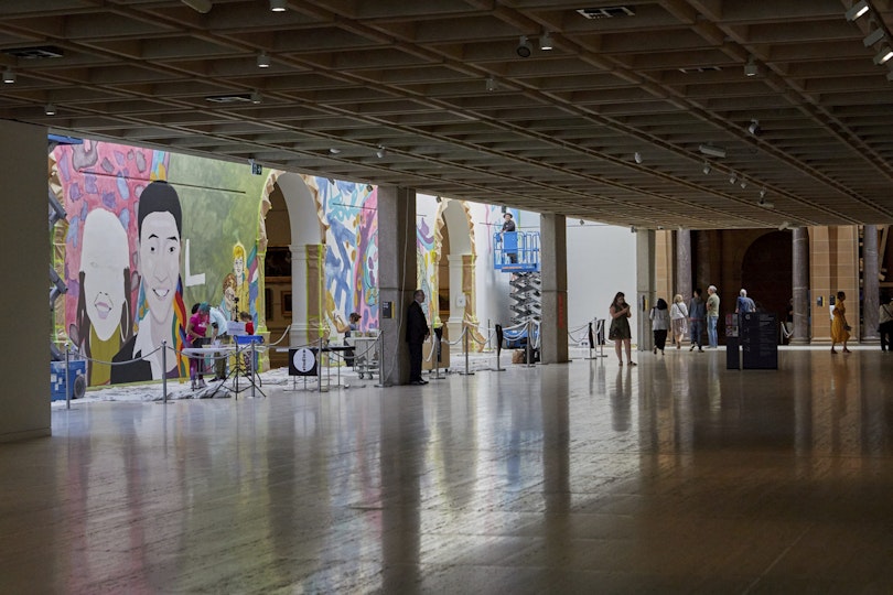 Parts of a colourful mural are visible looking across a  large, darkened gallery space.