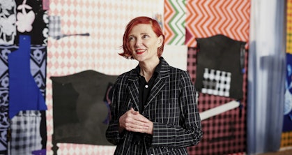 A woman with chin-length red hair, weaing a shirt and checked jacket, stands with her hands clasped in front of hanging textiles of various colours and patterns.