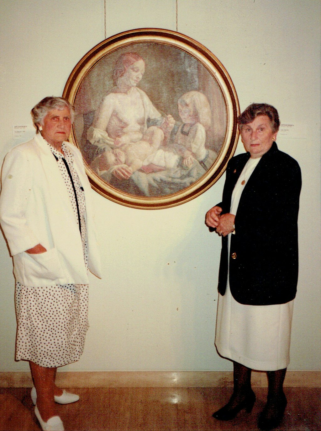 Two elderly women stand either side of a circular painting depicting a woman, a small child and a baby,